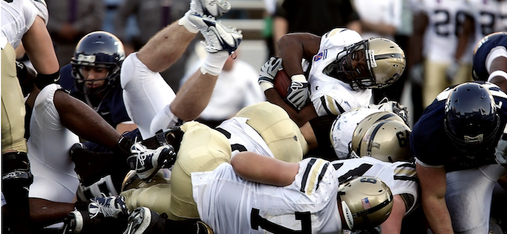 Six American football players in a pile. Some wear golden helmets, others wear black helmets. They are tackling someone.